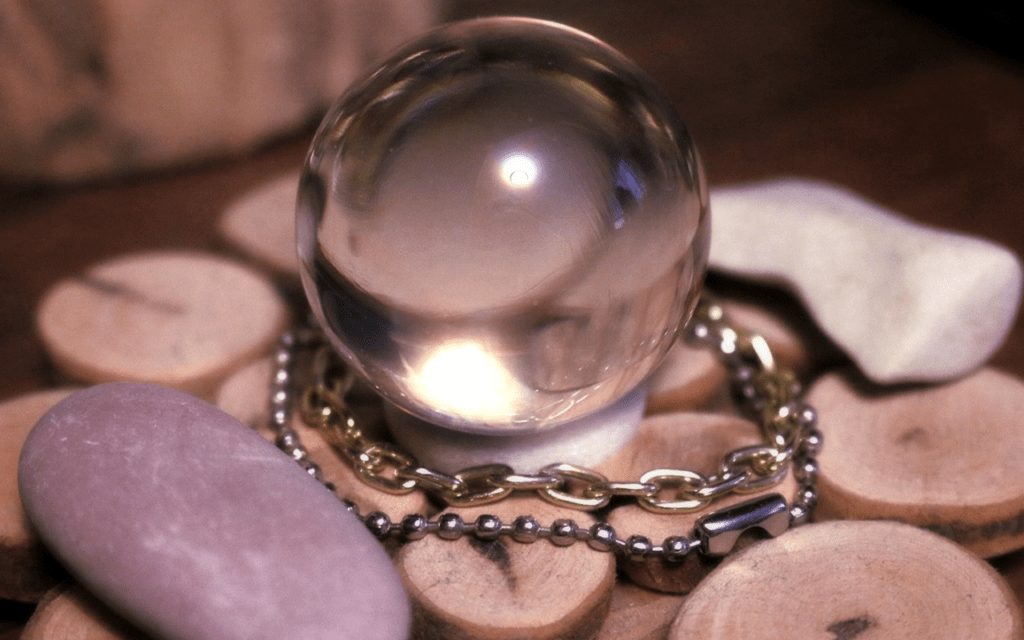 A clear crystal ball balanced on a pile of wooden coins and stones surrounded by beads and a chain.