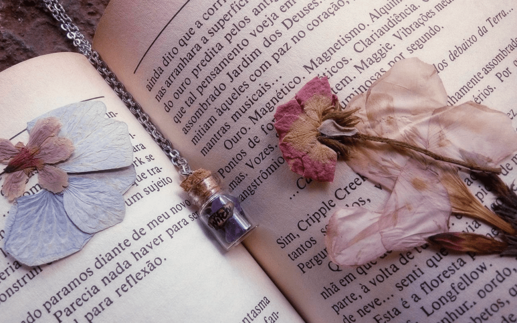 A close-up of an open novel with pressed flowers covering the pages and a small jar of amethyst chips on a chain as a bookmark.