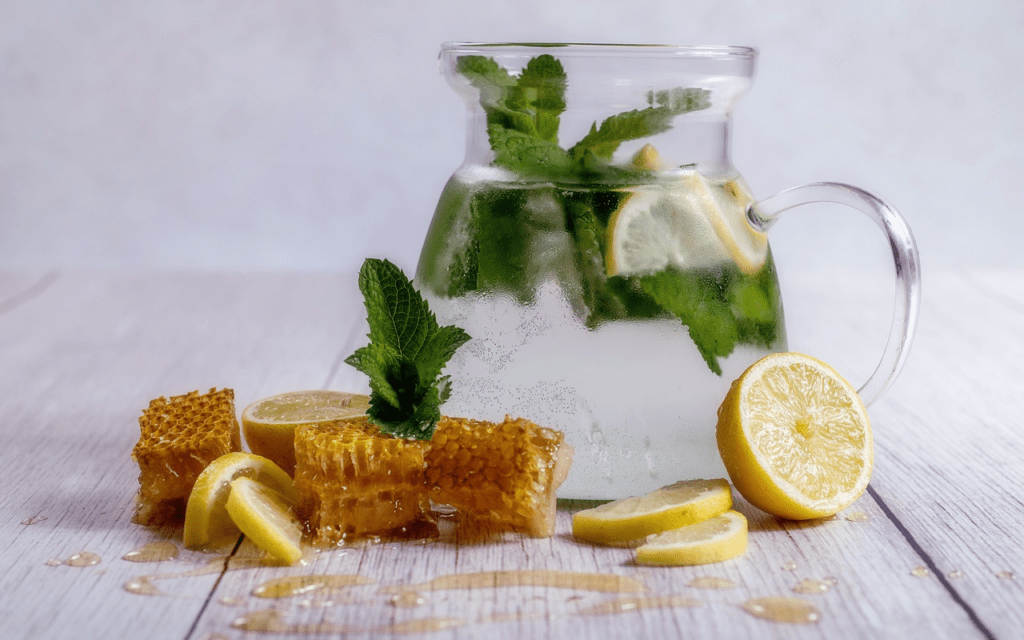 A glass pitcher of water with mint and lemon on a wooden table surrounded by lemon wedges, mint, and honeycomb.